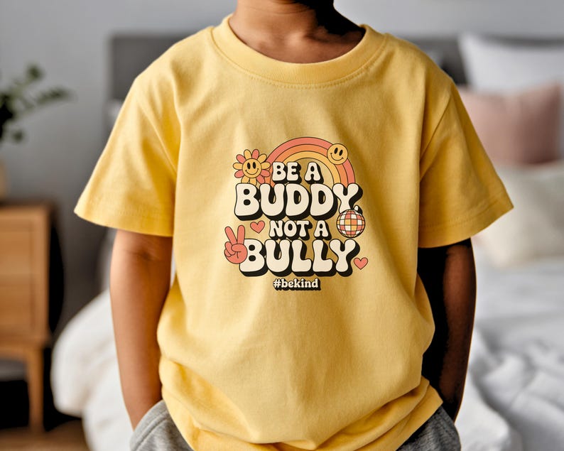 May include: A yellow t-shirt with the text "BE A BUDDY NOT A BULLY" in a retro font, featuring a rainbow, smiley face, flower, disco ball, and peace sign. The hashtag #bekind is also included.