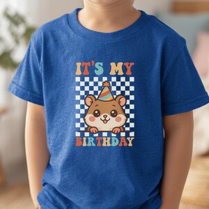 May include: A blue t-shirt featuring a cartoon hamster wearing a party hat, set against a checkered background. The text "IT'S MY BIRTHDAY" is displayed in colorful, retro-style lettering, perfect for a birthday celebration.