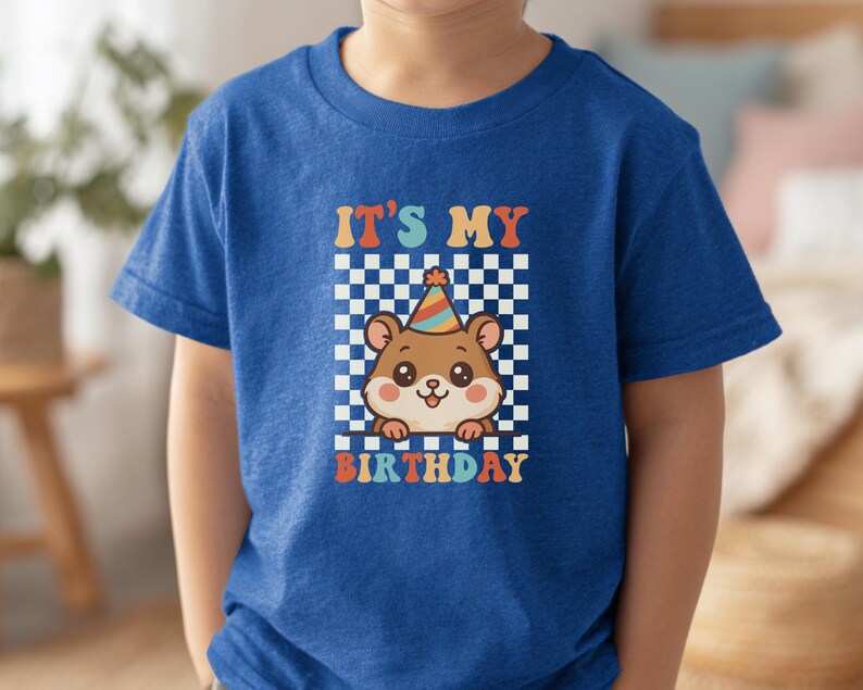 May include: A blue t-shirt featuring a cartoon hamster wearing a party hat, set against a checkered background. The text "IT'S MY BIRTHDAY" is displayed in colorful, retro-style lettering, perfect for a birthday celebration.