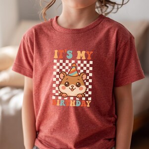 May include: A red t-shirt featuring a cartoon hamster wearing a party hat, set against a checkered background. The text "IT'S MY BIRTHDAY" is displayed above and below the hamster in retro-style lettering. The shirt is made of a soft material.