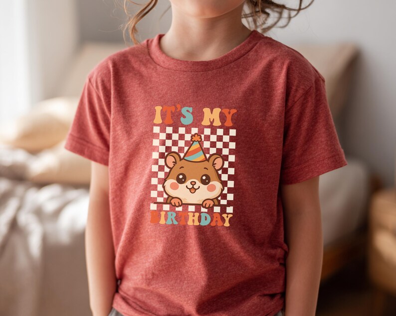 May include: A red t-shirt featuring a cartoon hamster wearing a party hat, set against a checkered background. The text "IT'S MY BIRTHDAY" is displayed above and below the hamster in retro-style lettering. The shirt is made of a soft material.