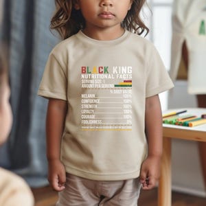 May include: A tan t-shirt with the text "BLACK KING NUTRITIONAL FACTS" in black and red. The shirt lists nutritional values for melanin, confidence, strength, loyalty, courage, and foolishness. The child is wearing khaki pants.