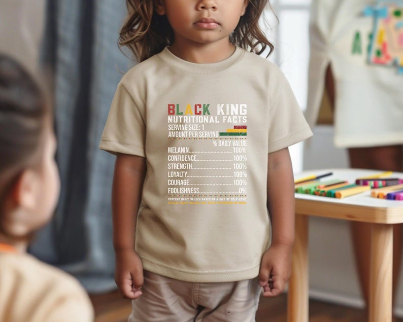 May include: A tan t-shirt with the text "BLACK KING NUTRITIONAL FACTS" in black and red. The shirt lists nutritional values for melanin, confidence, strength, loyalty, courage, and foolishness. The child is wearing khaki pants.