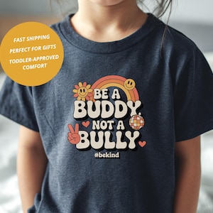 May include: A navy blue t-shirt with the text "BE A BUDDY NOT A BULLY" in a retro font, featuring a rainbow, flower, and peace sign graphic. The shirt also includes the hashtag #bekind.