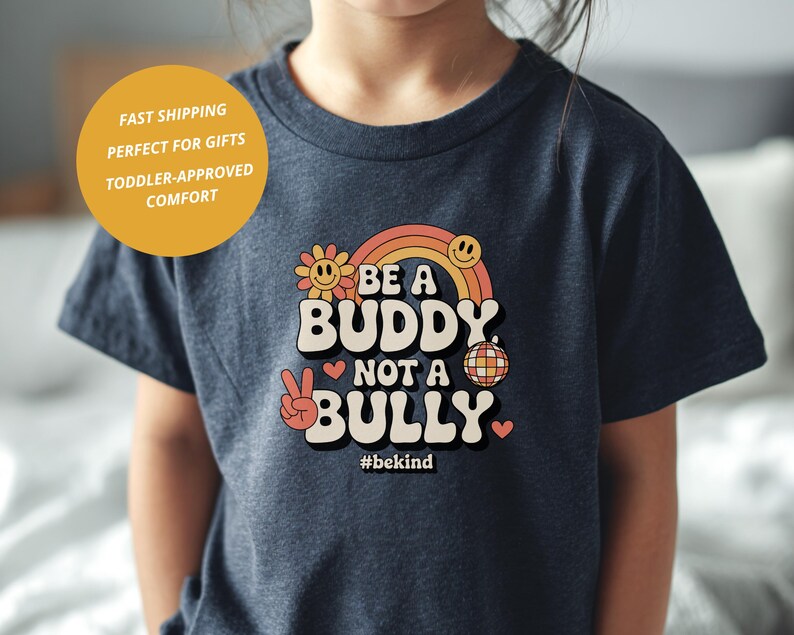 May include: A navy blue t-shirt with the text "BE A BUDDY NOT A BULLY" in a retro font, featuring a rainbow, flower, and peace sign graphic. The shirt also includes the hashtag #bekind.