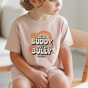 May include: A light pink t-shirt with the words "BE A BUDDY NOT A BULLY" in a retro font, with a rainbow, flower, peace sign, and disco ball graphic. The hashtag #bekind is below the text.