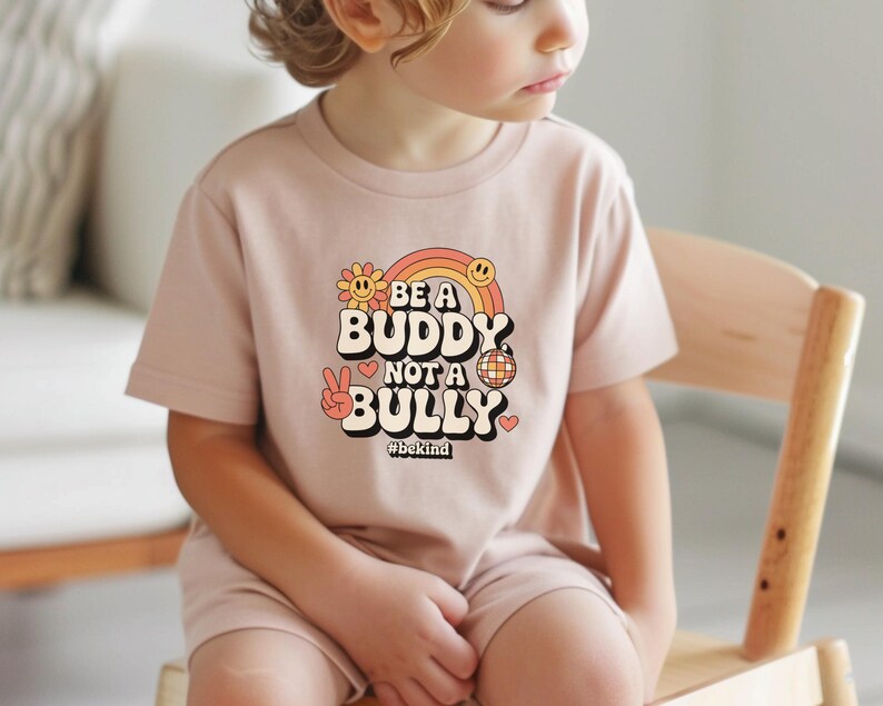 May include: A light pink t-shirt with the words "BE A BUDDY NOT A BULLY" in a retro font, with a rainbow, flower, peace sign, and disco ball graphic. The hashtag #bekind is below the text.