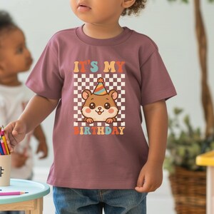 May include: A mauve-colored t-shirt featuring a cartoon hamster wearing a party hat, set against a checkered background. The text "IT'S MY BIRTHDAY" is displayed above and below the hamster illustration. The shirt is worn by a child.