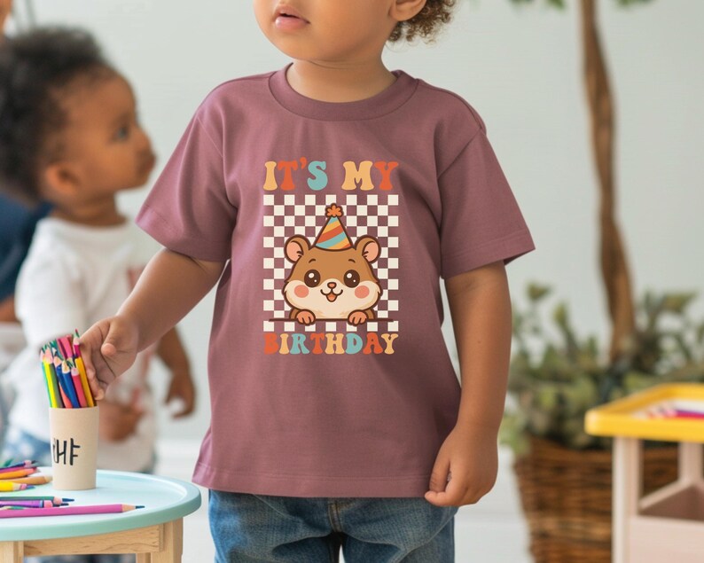 May include: A mauve-colored t-shirt featuring a cartoon hamster wearing a party hat, set against a checkered background. The text "IT'S MY BIRTHDAY" is displayed above and below the hamster illustration. The shirt is worn by a child.