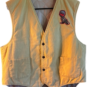 Puede incluir: Un chaleco acolchado de color beige con un parche bordado en rojo y negro de una mano sosteniendo una copa de vino. El chaleco tiene dos bolsillos y cierre de botones.