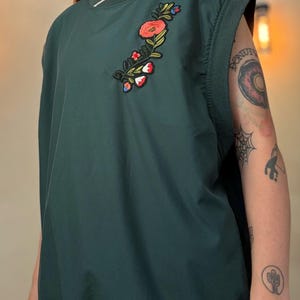 Puede incluir: Una camisa verde oscuro sin mangas con un diseño de bordado floral en el lado izquierdo del pecho. El bordado presenta flores rosas, rojas y azules.