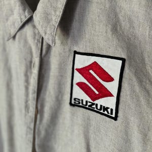 May include: A gray work shirt with a black and white embroidered patch that says "SUZUKI" in white letters with a red "S" logo.
