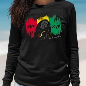 May include: Black long-sleeve shirt featuring a graphic of Bob Marley with the words "ONE LOVE" in red, yellow, and green brushstrokes. The shirt also includes the signature "bob marley" in white script.