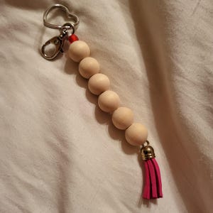 May include: A wooden bead keychain with a pink tassel. The keychain has a silver key ring and a red bead at the top.