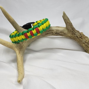 May include: A braided paracord bracelet in green, yellow, and red, secured with a black plastic buckle. The bracelet is displayed on a deer antler against a white background.