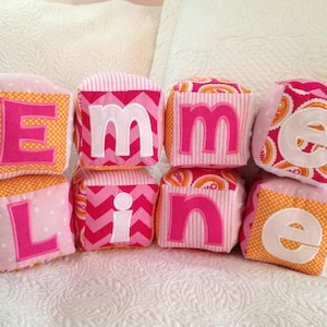 May include: Six fabric blocks spelling out the name "Emmeline" in pink and white letters. The blocks are made of different patterned fabrics in pink, orange, and white.