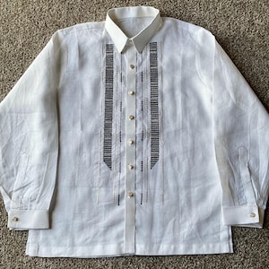 May include: White long-sleeved shirt with a classic collar and buttoned cuffs. The shirt features vertical embroidered panels in black and gold, with decorative buttons down the front. A formal garment.