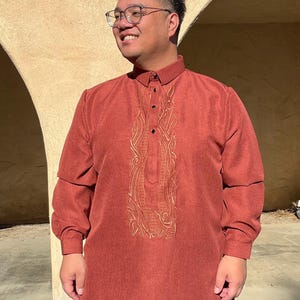 May include: A rust-coloured long-sleeved shirt with detailed gold embroidery down the front. The shirt features a mandarin collar and buttoned cuffs. The person is wearing blue trousers. The shirt is made of a textured fabric.