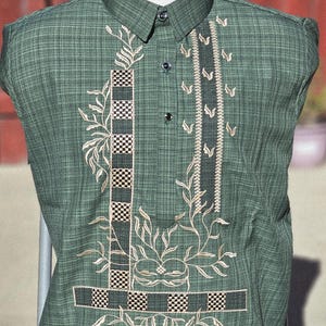 May include: A short-sleeved green shirt with a subtle grid pattern, featuring a button-up collar and a front placket. The fabric is decorated with intricate beige floral embroidery and dark, checkered geometric panels on the chest area.