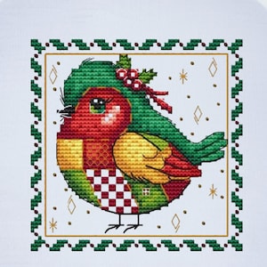 May include: A cross-stitch embroidery of a colorful bird inside a wooden hoop. The bird features red, green, yellow, and checkered patterns. The design is framed with a green border and gold accents, set against a white background.