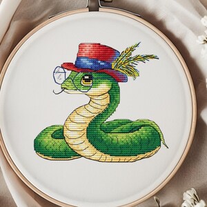 May include: A green snake wearing a red, blue, and white hat with a feather and glasses. The snake is embroidered on a white canvas in a wooden embroidery hoop.