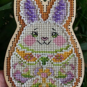 May include: Wooden bunny-shaped ornament with a cross-stitched design. The bunny features purple ears, pink cheeks, and a floral design in shades of pink, green, and yellow. The border is a light brown color, and the eyes are green.