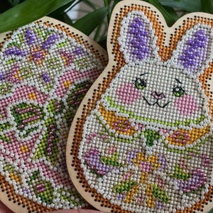 May include: Two wooden Easter ornaments, one shaped like an egg with a pink, green, and purple floral design, and the other shaped like a bunny with a white face and green, yellow, and purple floral design.