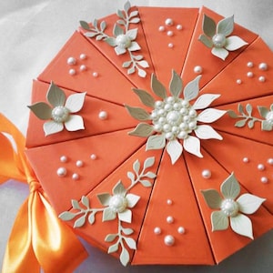 Könnte beinhalten: Orangefarbene, achteckige Geschenkbox mit dekorativem Blumendesign. Die Box ist in dreieckige Abschnitte unterteilt, die mit weißen Blumen, Perlenakzenten und Blätterzweigen verziert sind. Eine große orangefarbene Schleife ist an der Seite gebunden.