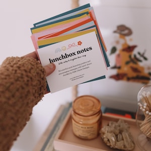 May include: A stack of colorful lunchbox notes with the text "lunchbox notes" and "little notes to brighten their day." The cards feature hand-drawn doodles and encouraging messages. The cards are held in a person's hand.
