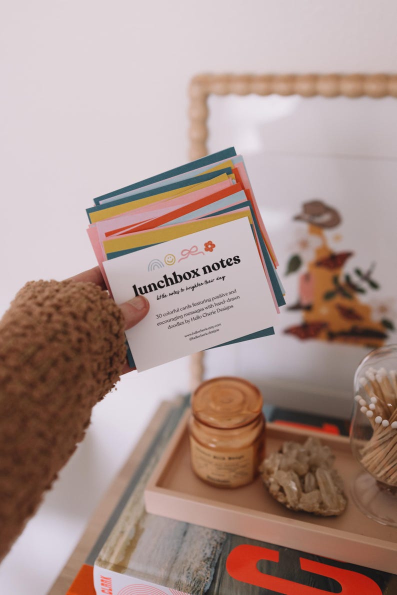 May include: A stack of colorful lunchbox notes with the text "lunchbox notes" and "little notes to brighten their day." The cards feature hand-drawn doodles and encouraging messages. The cards are held in a person's hand.