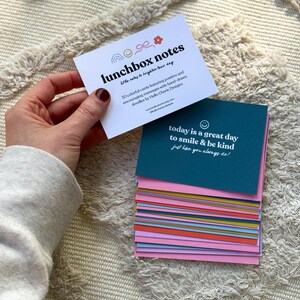 May include: A hand holds a white card with the text "lunchbox notes" and a stack of colorful cards. The top card reads "today is a great day to smile & be kind." The cards are designed to brighten the day with positive messages.