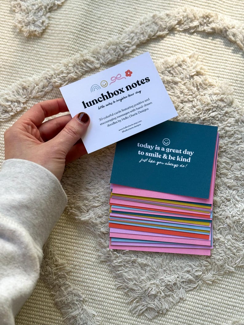 May include: A hand holds a white card with the text "lunchbox notes" and a stack of colorful cards. The top card reads "today is a great day to smile & be kind." The cards are designed to brighten the day with positive messages.