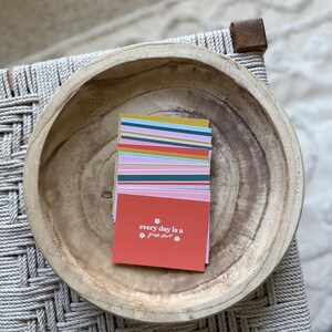 May include: A stack of colorful cards with the text "every day is a fresh start" in a wooden bowl. The cards have stripes of various colors, including pink, blue, and yellow. The bowl is light brown and sits on a woven surface.