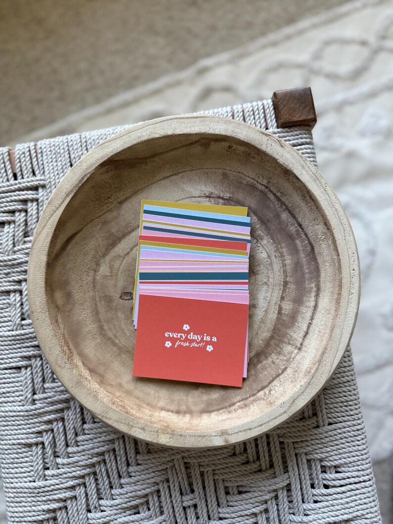 May include: A stack of colorful cards with the text "every day is a fresh start" in a wooden bowl. The cards have stripes of various colors, including pink, blue, and yellow. The bowl is light brown and sits on a woven surface.