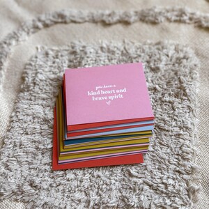May include: A stack of colorful square cards with the text "you have a kind heart and brave spirit" on the top card. The cards are stacked on a textured, off-white surface.