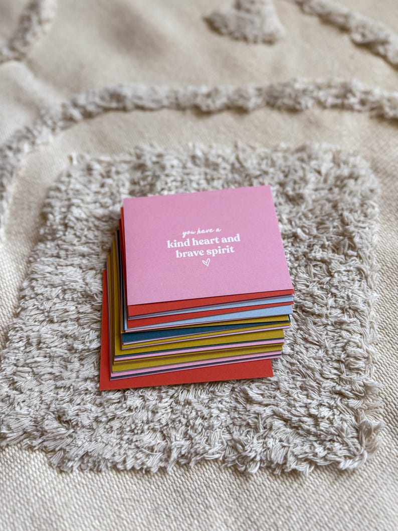 May include: A stack of colorful square cards with the text "you have a kind heart and brave spirit" on the top card. The cards are stacked on a textured, off-white surface.