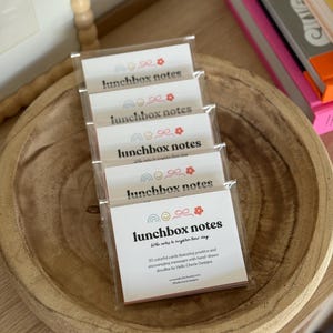May include: A stack of packaged lunchbox notes in a wooden bowl. Each package contains cards with the text "lunchbox notes" and a rainbow, bow, and flower design. The text "Little notes to brighten their day" is also visible.