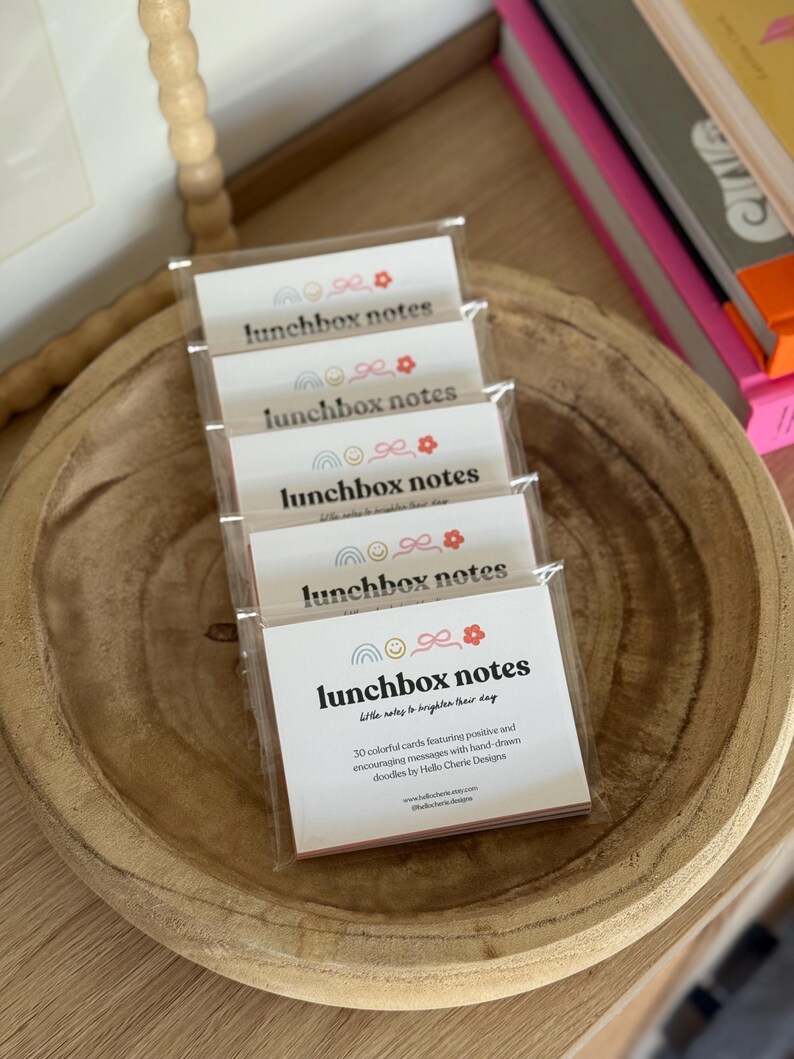 May include: A stack of packaged lunchbox notes in a wooden bowl. Each package contains cards with the text "lunchbox notes" and a rainbow, bow, and flower design. The text "Little notes to brighten their day" is also visible.