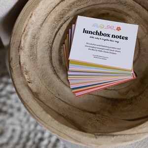 May include: A stack of colorful lunchbox notes in a wooden bowl. The notes have a white background with the text "lunchbox notes" and "Little notes to brighten their day". Each card features a rainbow, bow, and flower.