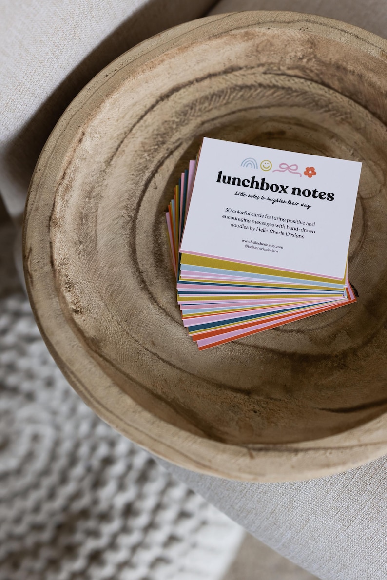 May include: A stack of colorful lunchbox notes in a wooden bowl. The notes have a white background with the text "lunchbox notes" and "Little notes to brighten their day". Each card features a rainbow, bow, and flower.
