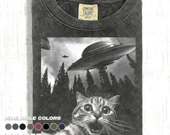 May include: Dark gray t-shirt with a graphic of a cat looking up at a UFO. The cat has a surprised expression. The shirt has the text "COMFORT COLORS 1717" on the tag. The words "AVAILABLE COLORS" and "PEPPER" are also visible.