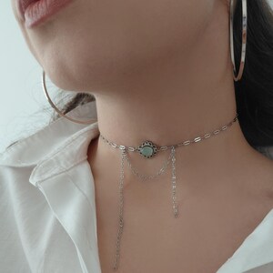 Puede incluir: Un collar gargantilla plateado con un dise&ntilde;o floral central y una piedra verde claro. El collar tiene delicadas cadenas colgantes. Tambi&eacute;n se ven pendientes de aro plateados grandes.