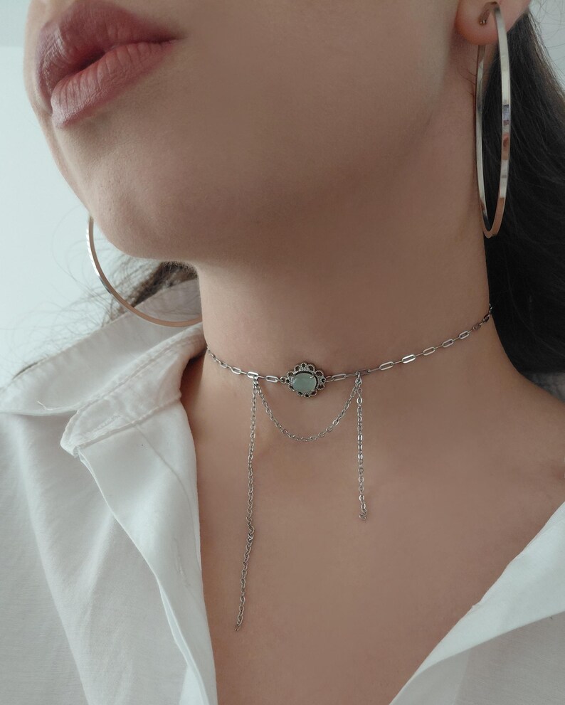 Puede incluir: Un collar gargantilla plateado con un dise&ntilde;o floral central y una piedra verde claro. El collar tiene delicadas cadenas colgantes. Tambi&eacute;n se ven pendientes de aro plateados grandes.