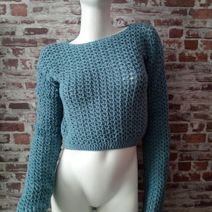 Peut inclure: Un pull court en crochet de couleur sarcelle avec des manches longues. Le pull a une encolure large et un motif de point texturé. Le vêtement est présenté sur un mannequin blanc devant un mur de briques.