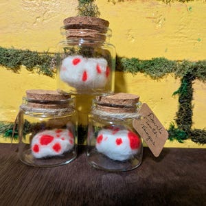 May include: Three small glass jars with cork lids, each containing a white, fluffy object dotted with red spots. A small tag reads "Devil's Tooth Fungus". The jars are arranged in a small stack.