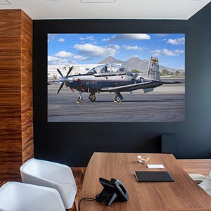 May include: A framed print of a U.S. Air Force T-6 Texan II aircraft on a runway. The plane is dark blue and white with a propeller and the words "U.S. AIR FORCE" on the side. The background shows mountains and a blue sky with clouds.