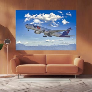 May include: A large framed print of a FedEx cargo plane taking off against a blue sky with fluffy white clouds. The plane is white with red and purple accents. The print hangs above an orange sofa.