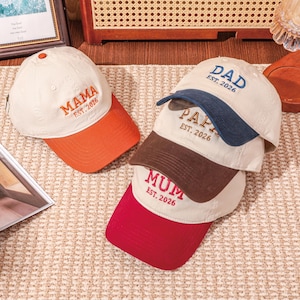 May include: A collection of baseball caps with embroidered text. The caps are cream-coloured with coloured brims in orange, navy, brown, and red. The text on the caps reads "MAMA", "DAD", "PAP", and "MUM", each followed by "EST. 2026".