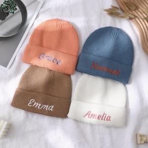 May include: Four ribbed knit beanies in peach, blue, brown, and white. Each beanie has a name embroidered in a contrasting color: "Grace", "Hannah", "Emma", and "Amelia". The beanies are arranged on a white surface.