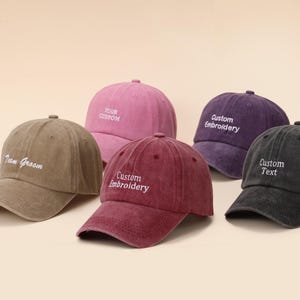 Peut inclure: Cinq casquettes de baseball de couleurs variées : marron, rose, bordeaux, violet et noir. Chaque casquette présente une broderie personnalisée blanche avec des phrases comme "Team Groom" et "Your Custom".